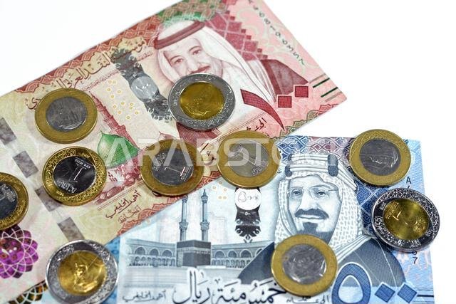 مجموعة من الأوراق النقدية السعودية من فئتي 500 و100 ريال سعودي، بالإضافة إلى كومة من العملات المعدنية السعودية من فئة ريال واحد وريالين، تحمل صورة الملك سلمان بن عبد العزيز.