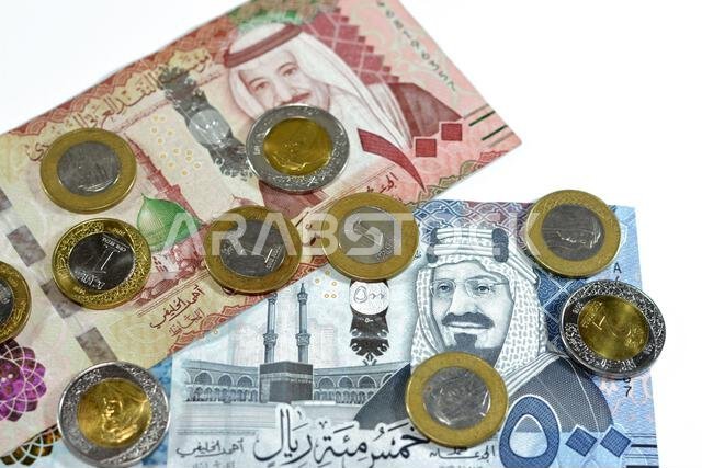 مجموعة من الأوراق النقدية السعودية من فئتي 500 و100 ريال سعودي، بالإضافة إلى كومة من العملات المعدنية السعودية من فئة ريال واحد وريالين، تحمل صورة الملك سلمان بن عبد العزيز.