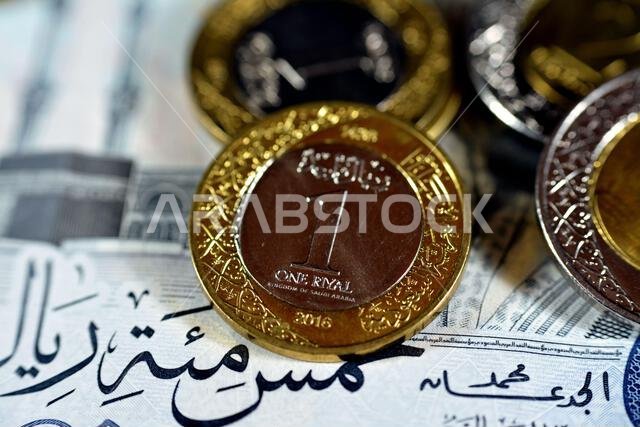 الوجه الخلفي لعملة معدنية من فئة ريال سعودي واحد (1 ريال) من سلسلة 1438 هـ 2016، معزولة مع عملات سعودية أخرى على ورقة نقدية من فئة خمسمائة ريال (500 ريال).