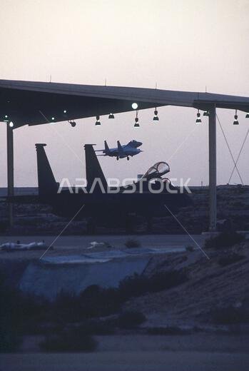 An F-15 Eagle aircraft of the 1ST Tactical Fighter Wing sits in a hangar as another F-15 takes off during Operation Desert Shield. Subject Operation/Series: DESERT SHIELD Country: Saudi Arabia (SAU)