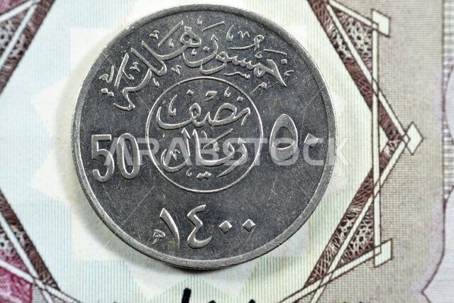 الوجه الخلفي لعملة نصف ريال سعودية قديمة من فئة 50 هللة، ترجمة (50 هللة، سلسلة عملات نصف ريال، 1400 هـ)، نقش فوق الكتابة داخل دائرة تقسم حرف v الوجه الخلفي لعملة نصف ريال سعودية قديمة من فئة 50 هللة، ترجمة (50 هللة، سلسلة عملات نصف ريال، 1400 هـ)، نقش فوق الكتابة داخل دائرة تقسم حرف v