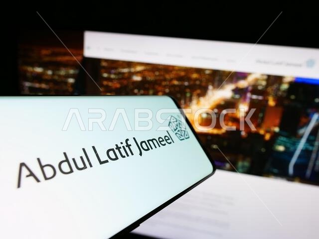 Mobile phone with logo of Saudi Arabian company Abdul Latif Jameel (ALJ) on screen in front of website. Focus on center-right of phone display. Mobile phone with logo of Saudi Arabian company Abdul Latif Jameel (ALJ) on screen in front of website. Focus on center-right of phone display.
