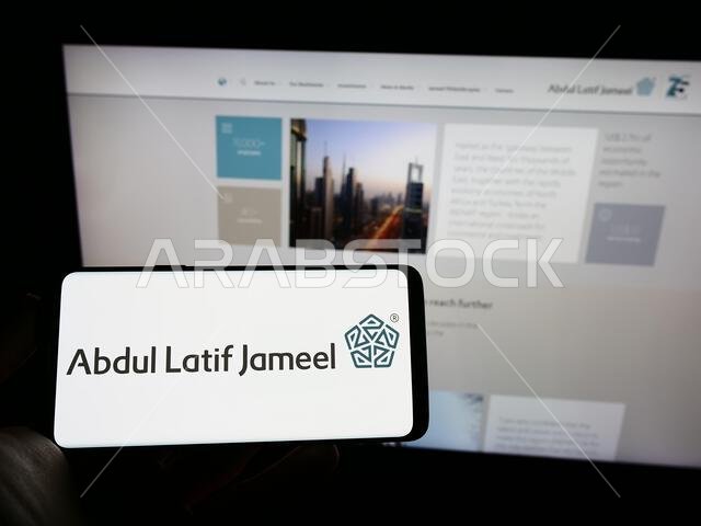 شخص يحمل هاتفًا محمولًا عليه شعار شركة عبد اللطيف جميل السعودية (ALJ) على الشاشة أمام صفحة الويب. التركيز على شاشة الهاتف. شخص يحمل هاتفًا محمولًا عليه شعار شركة عبد اللطيف جميل السعودية (ALJ) على الشاشة أمام صفحة الويب. التركيز على شاشة الهاتف.