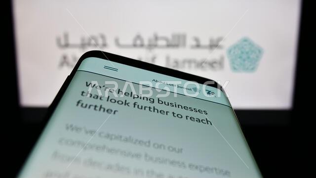 هاتف ذكي يعرض موقع شركة عبد اللطيف جميل (ALJ) السعودية على الشاشة أمام شعار الشركة. التركيز على الزاوية العلوية اليسرى من شاشة الهاتف.