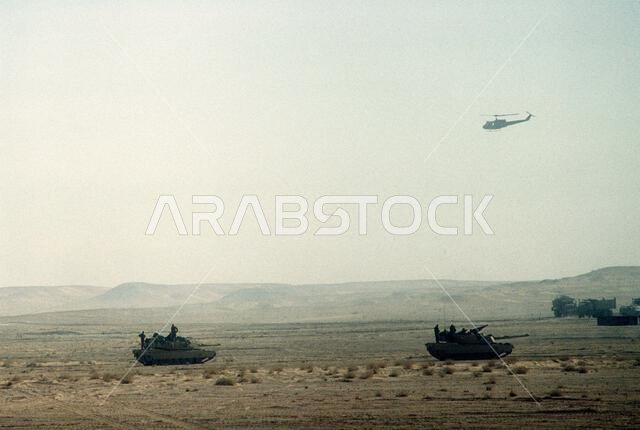 A UH-1 Iroquois helicopter flies overhead as M-1A1 Abrams main battle tanks of Co. A, 3rd Bn., 32nd Armored Regt., 1ST Cav. Div., move out on an exercise during Operation Desert Shield. Subject Operation/Series: DESERT SHIELD Country: Saudi Arabia(SAU) A UH-1 Iroquois helicopter flies overhead as M-1A1 Abrams main battle tanks of Co. A, 3rd Bn., 32nd Armored Regt., 1ST Cav. Div., move out on an exercise during Operation Desert Shield. Subject Operation/Series: DESERT SHIELD Country: Saudi Arabia(SAU)