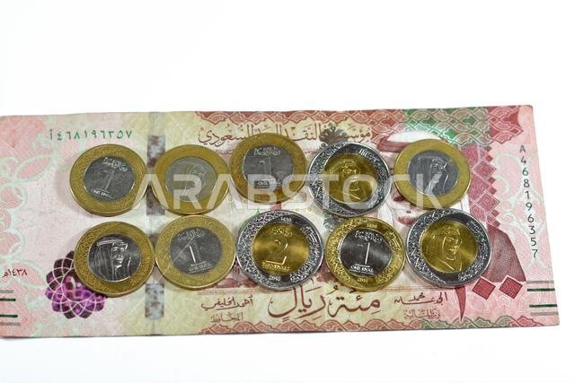 ورقة نقدية من فئة 100 ريال سعودي مع رزمة من العملات المعدنية من فئة ريال واحد وريالين، تحمل صورة الملك سلمان بن عبد العزيز آل سعود.