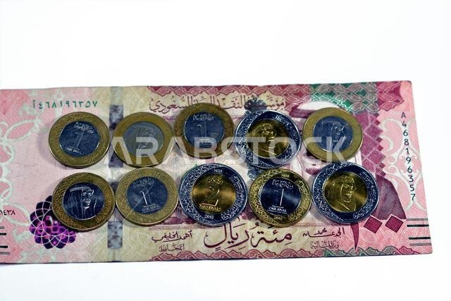 ورقة نقدية من فئة 100 ريال سعودي مع رزمة من العملات المعدنية من فئة ريال واحد وريالين، تحمل صورة الملك سلمان بن عبد العزيز آل سعود.
