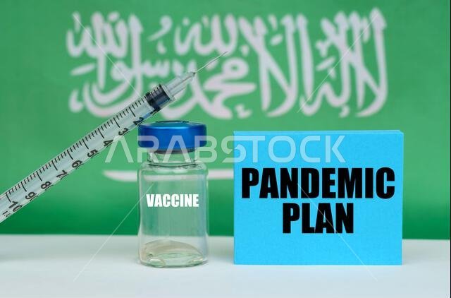 مفهوم طبي. لقاح، حقنة، ولوحة زرقاء منقوش عليها عبارة "خطة لمواجهة الجائحة". في الخلفية علم المملكة العربية السعودية