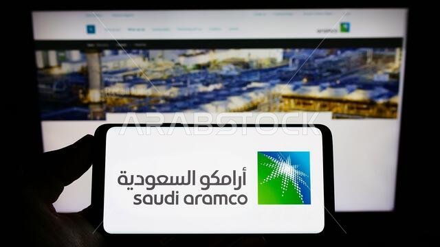 Person holding smartphone with logo of oil and gas company Saudi Aramco on screen in front of website. Focus on phone display. Person holding smartphone with logo of oil and gas company Saudi Aramco on screen in front of website. Focus on phone display.