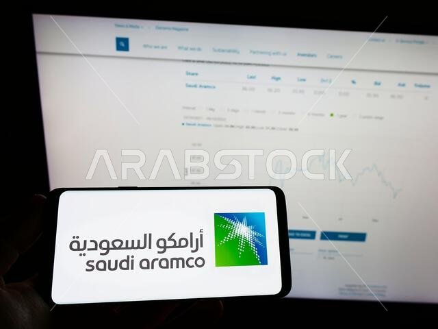 شخص يحمل هاتفًا محمولًا عليه شعار شركة النفط والغاز السعودية أرامكو السعودية على الشاشة أمام صفحة ويب خاصة بالشركة. التركيز على شاشة الهاتف.