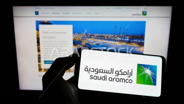 شخص يحمل هاتفًا محمولًا عليه شعار شركة النفط والغاز السعودية أرامكو السعودية على الشاشة أمام صفحة ويب خاصة بالشركة. التركيز على شاشة الهاتف. شخص يحمل هاتفًا محمولًا عليه شعار شركة النفط والغاز السعودية أرامكو السعودية على الشاشة أمام صفحة ويب خاصة بالشركة. التركيز على شاشة الهاتف.