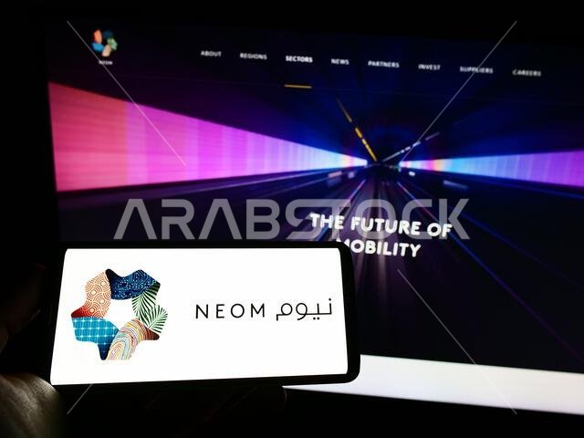 شخص يحمل هاتفًا محمولًا عليه شعار شركة نيوم العقارية السعودية على الشاشة أمام صفحة ويب. التركيز على شاشة الهاتف.