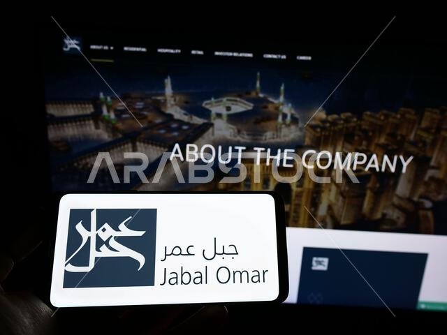 شخص يحمل هاتفًا محمولًا عليه شعار شركة جبل عمر للتطوير (ش.م.ع.) على الشاشة أمام صفحة الويب الخاصة بالشركة. التركيز على شاشة الهاتف.