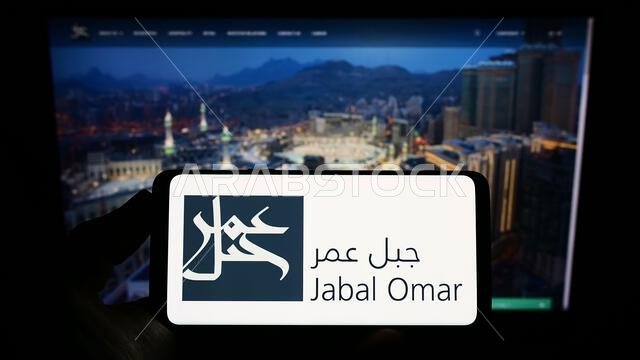 شخص يحمل هاتفًا ذكيًا عليه شعار شركة جبل عمر للتطوير (ش.م.ع.) على الشاشة أمام موقع إلكتروني. التركيز على شاشة الهاتف.