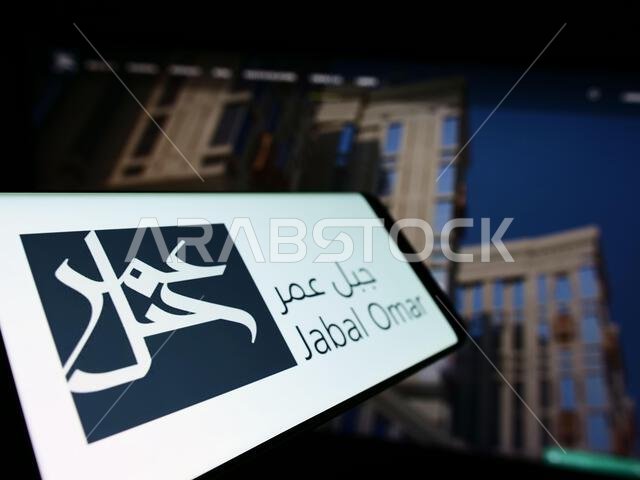 هاتف ذكي يعرض شعار شركة جبل عمر للتطوير (ش.م.ع) على شاشته أمام موقع الشركة الإلكتروني. التركيز على يسار شاشة الهاتف. هاتف ذكي يعرض شعار شركة جبل عمر للتطوير (ش.م.ع) على شاشته أمام موقع الشركة الإلكتروني. التركيز على يسار شاشة الهاتف.