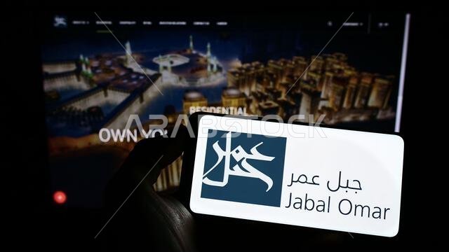 شخص يحمل هاتفًا محمولًا عليه شعار شركة جبل عمر للتطوير (ش.م.ع.) على الشاشة أمام صفحة الويب الخاصة بالشركة. التركيز على شاشة الهاتف.