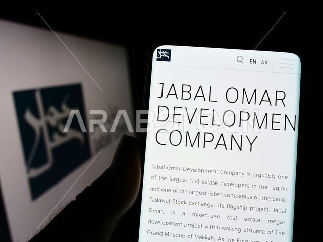 شخص يحمل هاتفًا محمولًا يعرض صفحة ويب لشركة جبل عمر للتطوير (ش.م.ع.) أمام الشعار. التركيز على منتصف شاشة الهاتف.