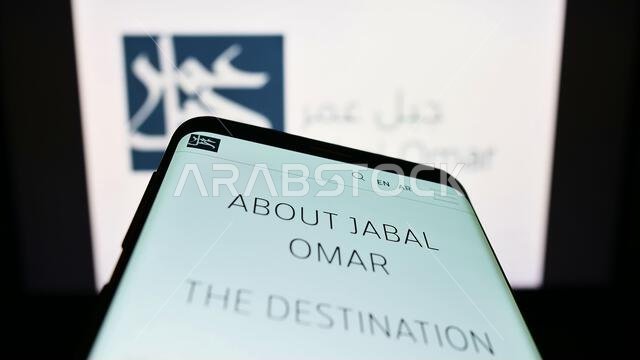 هاتف محمول يعرض موقع شركة جبل عمر للتطوير (ش.م.ع) أمام شعار الشركة. التركيز على أعلى يسار شاشة الهاتف. هاتف محمول يعرض موقع شركة جبل عمر للتطوير (ش.م.ع) أمام شعار الشركة. التركيز على أعلى يسار شاشة الهاتف.