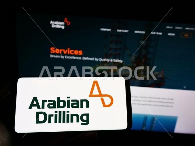 شخص يحمل هاتفًا محمولًا عليه شعار شركة الحفر العربية السعودية (ADC) على الشاشة أمام صفحة الويب. التركيز على شاشة الهاتف. شخص يحمل هاتفًا محمولًا عليه شعار شركة الحفر العربية السعودية (ADC) على الشاشة أمام صفحة الويب. التركيز على شاشة الهاتف.