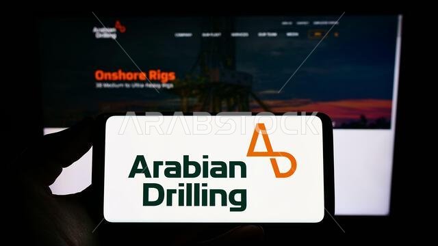 Person holding mobile phone with logo of Saudi business Arabian Drilling Company (ADC) on screen in front of web page. Focus on phone display. Person holding mobile phone with logo of Saudi business Arabian Drilling Company (ADC) on screen in front of web page. Focus on phone display.