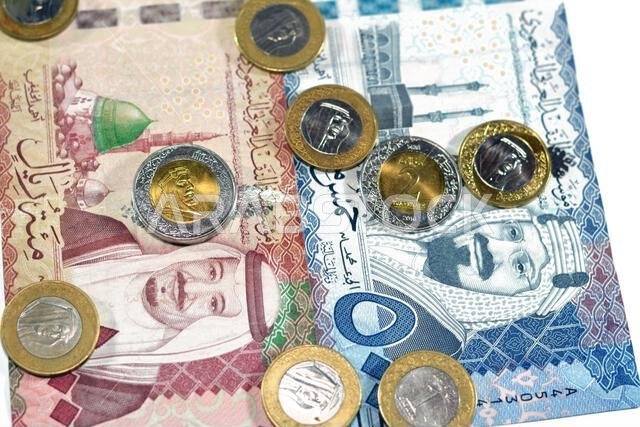 مجموعة من الأوراق النقدية السعودية من فئتي 500 و100 ريال سعودي، بالإضافة إلى كومة من العملات المعدنية السعودية من فئة ريال واحد وريالين، تحمل صورة الملك سلمان بن عبد العزيز.