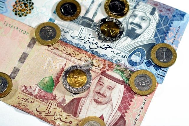 مجموعة من الأوراق النقدية السعودية من فئتي 500 و100 ريال سعودي، بالإضافة إلى كومة من العملات المعدنية السعودية من فئة ريال واحد وريالين، تحمل صورة الملك سلمان بن عبد العزيز.