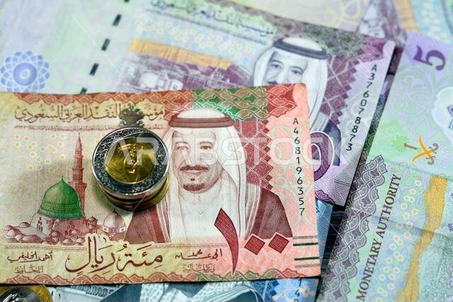 أوراق نقدية وعملات معدنية من الريال السعودي، تتضمن رزمة من فئات 500 و100 و5 ريالات، بالإضافة إلى عملات معدنية من فئتي 1 و2 ريال، تحمل صورة الملك سلمان بن عبد العزيز آل سعود.