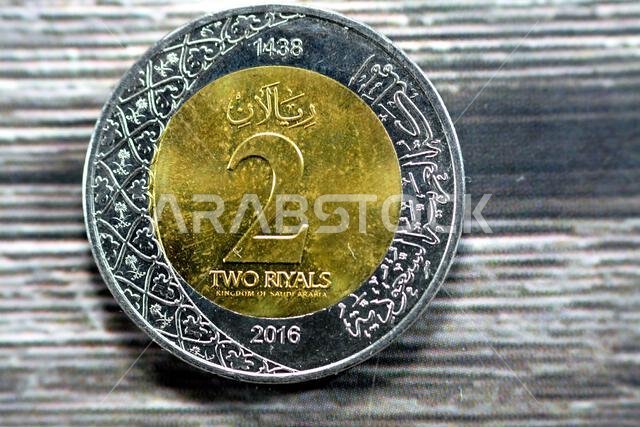 الوجه الخلفي لعملة معدنية من فئة ريالين سعوديين، إصدار عام 1438 هـ/2016م، يحمل شعارات نبالة متعددة ضمن الزخارف النباتية ونقشًا لـ الوجه الخلفي لعملة معدنية من فئة ريالين سعوديين، إصدار عام 1438 هـ/2016م، يحمل شعارات نبالة متعددة ضمن الزخارف النباتية ونقشًا لـ