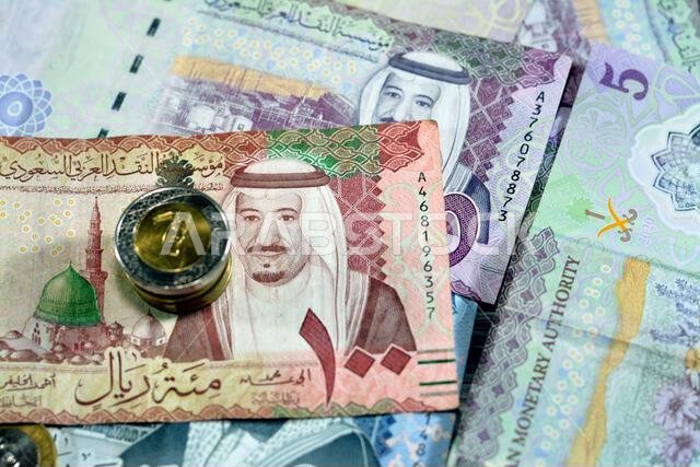 أوراق نقدية وعملات معدنية من الريال السعودي، تتضمن رزمة من فئات 500 و100 و5 ريالات، بالإضافة إلى عملات معدنية من فئتي 1 و2 ريال، تحمل صورة الملك سلمان بن عبد العزيز آل سعود.
