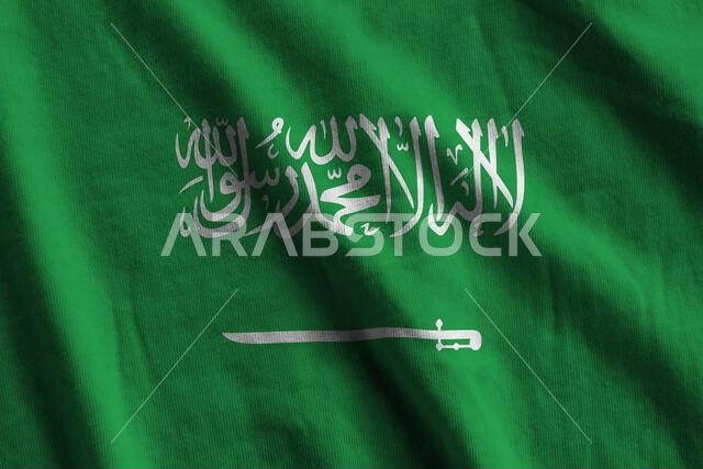علم المملكة العربية السعودية ذو الطيات الكبيرة يرفرف عن قرب تحت إضاءة الاستوديو الداخلية. الرموز والألوان الرسمية على لافتة قماشية