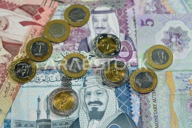 أوراق نقدية وعملات معدنية من الريال السعودي، تتضمن رزمة من فئات 500 و100 و5 ريالات، بالإضافة إلى عملات معدنية من فئتي 1 و2 ريال، تحمل صورة الملك سلمان بن عبد العزيز آل سعود. أوراق نقدية وعملات معدنية من الريال السعودي، تتضمن رزمة من فئات 500 و100 و5 ريالات، بالإضافة إلى عملات معدنية من فئتي 1 و2 ريال، تحمل صورة الملك سلمان بن عبد العزيز آل سعود.