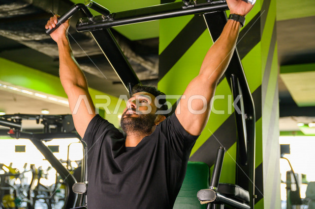 شاب عربي خليجي سعودي رياضي يقوم بتمرين العقلة السحب للأعلى ، تقوية العضلات ، كمال الأجسام و اللياقة البدنية ، ممارسة التمارين الرياضية في النادي الرياضي