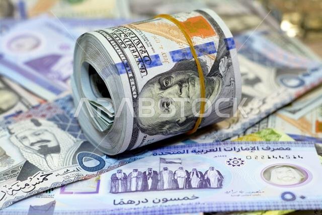 لفة نقود أمريكية عليها شريط، وكومة من أوراق الدولار الأمريكي، وأوراق نقدية أمريكية مع أوراق نقدية من الريال السعودي والإمارات الجديدة.