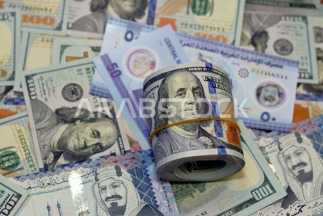 لفة نقود أمريكية عليها شريط، وكومة من أوراق الدولار الأمريكي، وأوراق نقدية أمريكية مع أوراق نقدية من الريال السعودي والإمارات الجديدة.