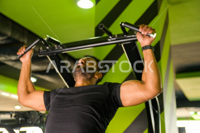 شاب عربي خليجي سعودي رياضي يقوم بتمرين العقلة السحب للأعلى ، تقوية العضلات ، كمال الأجسام و اللياقة البدنية ، ممارسة التمارين الرياضية في النادي الرياضي