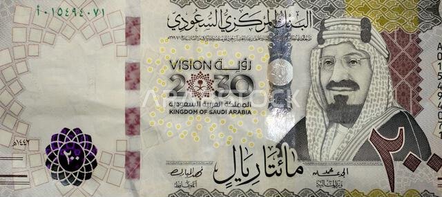 يظهر جزء كبير من الوجه الأمامي لورقة نقدية من فئة 200 ريال سعودي صورة الملك عبد العزيز آل سعود، مؤسس المملكة، وشعار ثلاثي الأبعاد.