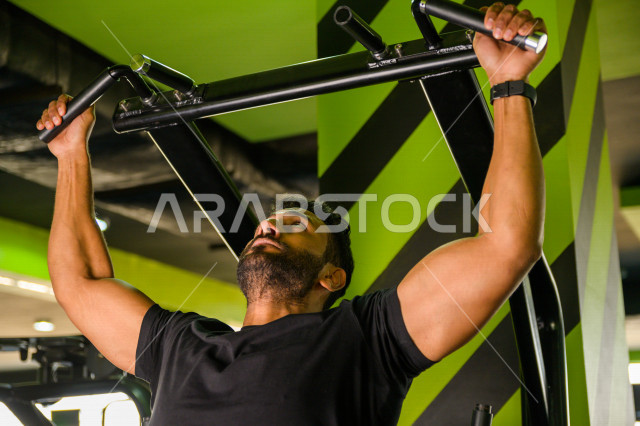 شاب عربي خليجي سعودي رياضي يقوم بتمرين العقلة السحب للأعلى ، تقوية العضلات ، كمال الأجسام و اللياقة البدنية ، ممارسة التمارين الرياضية في النادي الرياضي