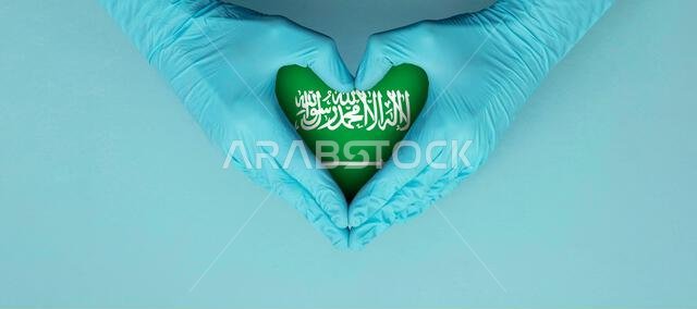 أيدي أطباء يرتدون قفازات جراحية زرقاء تشكل رمز القلب مع علم المملكة العربية السعودية