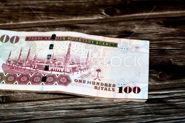 الوجه الخلفي لورقة نقدية سعودية من فئة 100 ريال سعودي، تحمل صورة المسجد النبوي الشريف في المدينة المنورة، معزولة على خلفية خشبية.