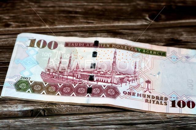 الوجه الخلفي لورقة نقدية سعودية من فئة 100 ريال سعودي، تحمل صورة المسجد النبوي الشريف في المدينة المنورة، معزولة على خلفية خشبية. الوجه الخلفي لورقة نقدية سعودية من فئة 100 ريال سعودي، تحمل صورة المسجد النبوي الشريف في المدينة المنورة، معزولة على خلفية خشبية.