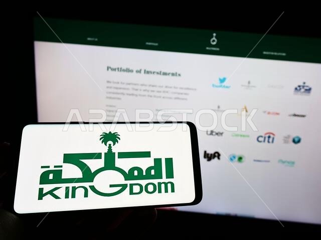 شخص يحمل هاتفًا محمولًا عليه شعار شركة المملكة العربية السعودية القابضة (KHC) على الشاشة أمام صفحة الويب. التركيز على شاشة الهاتف. شخص يحمل هاتفًا محمولًا عليه شعار شركة المملكة العربية السعودية القابضة (KHC) على الشاشة أمام صفحة الويب. التركيز على شاشة الهاتف.