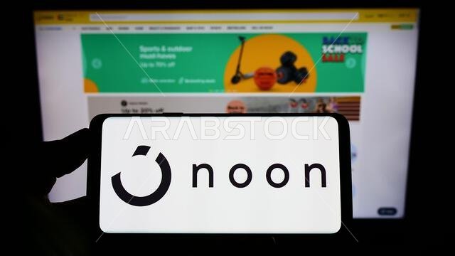شخص يحمل هاتفًا ذكيًا عليه شعار شركة التجارة الإلكترونية "نون إيه دي هولدينغز ليمتد" معروضًا على الشاشة أمام موقع إلكتروني. التركيز على شاشة الهاتف.