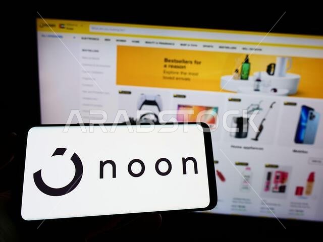 Person holding mobile phone with logo of e-commerce company Noon AD Holdings Ltd. on screen in front of business web page. Focus on phone display.