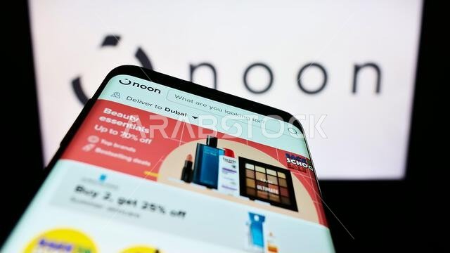 شخص يحمل هاتفًا محمولًا يعرض صفحة ويب لشركة التجارة الإلكترونية "نون إيه دي هولدينغز ليمتد" أمام الشعار. التركيز على أعلى يسار شاشة الهاتف.
