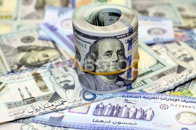 American money roll with a band on a pile of American dollar bills American cash money banknotes with Saudi Arabia riyals money banknotes and New Emir