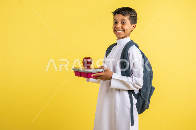 بورتريه لطالب عربي خليجي سعودي، يحمل الحقيبة المدرسية وبيده حافظة الطعام المدرسية وفاكهة التفاح الأحمر، فطور صباحي صحي، يرتدي الزي السعودي التقليدي، الإستعداد للعام الدراسي الجديد، التعليم الدراسي في السعودية، العودة إلى المدارس، خلفية صفراء