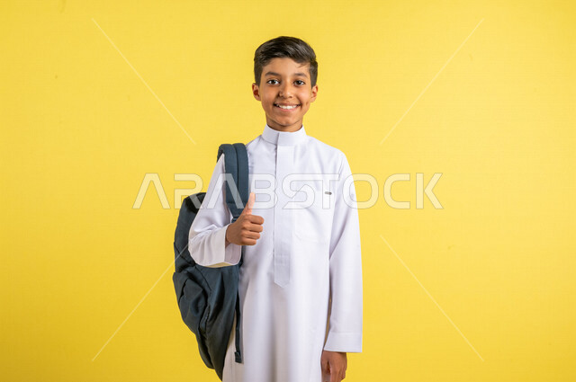 بورتريه لطالب عربي خليجي سعودي مبتسم ، يحمل الحقيبة المدرسية، يرتدي الزي السعودي التقليدي ، الإستعداد للعام الدراسي الجديد ، مرحلة دراسية ، التعليم الدراسي في المملكة العربية السعودية ، خلفية صفراء ، العودة إلى المدارس