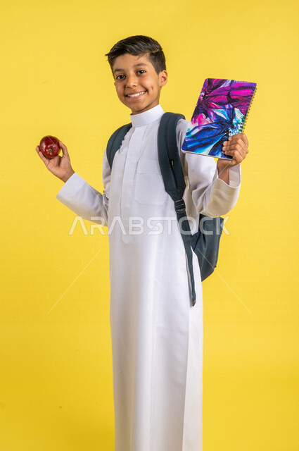 بورتريه لطالب عربي خليجي سعودي، يحمل الحقيبة المدرسية وبيده الدفتر المدرسي وفاكهة التفاح الأحمر، فطور صباحي صحي، يرتدي الزي السعودي التقليدي، الإستعداد للعام الدراسي الجديد، التعليم الدراسي في المملكة العربية السعودية، العودة إلى المدارس، خلفية صفراء