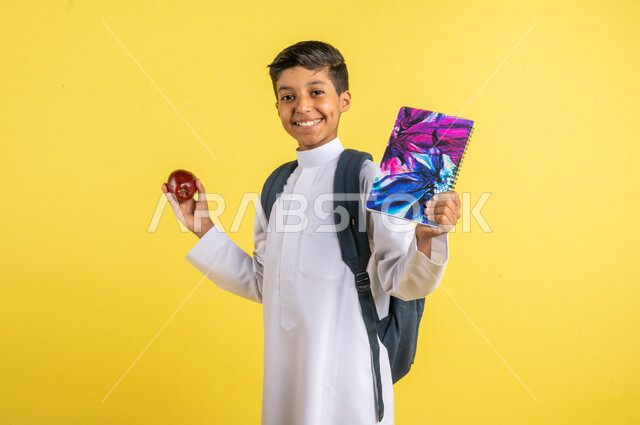 بورتريه لطالب عربي خليجي سعودي، يحمل الحقيبة المدرسية وبيده الدفتر المدرسي وفاكهة التفاح الأحمر، فطور صباحي صحي، يرتدي الزي السعودي التقليدي، الإستعداد للعام الدراسي الجديد، التعليم الدراسي في المملكة العربية السعودية، العودة إلى المدارس، خلفية صفراء