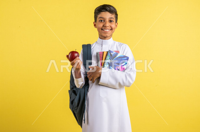 بورتريه لطالب عربي خليجي سعودي، يحمل الحقيبة المدرسية وبيده الدفاتر المدرسية وفاكهة التفاح الأحمر، فطور صباحي صحي، يرتدي الزي السعودي التقليدي، الإستعداد للعام الدراسي الجديد، التعليم الدراسي في المملكة العربية السعودية، العودة إلى المدارس، خلفية صفراء